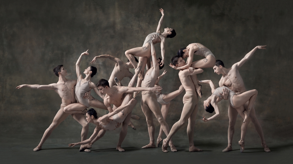 A group of ballet dancers in beige costumes form a dynamic, intertwined sculpture-like pose against a textured green background.
