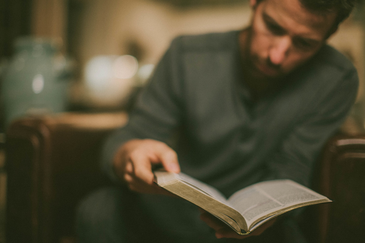 A man in a dimly lit room intently reads a book, holding it open in one hand.