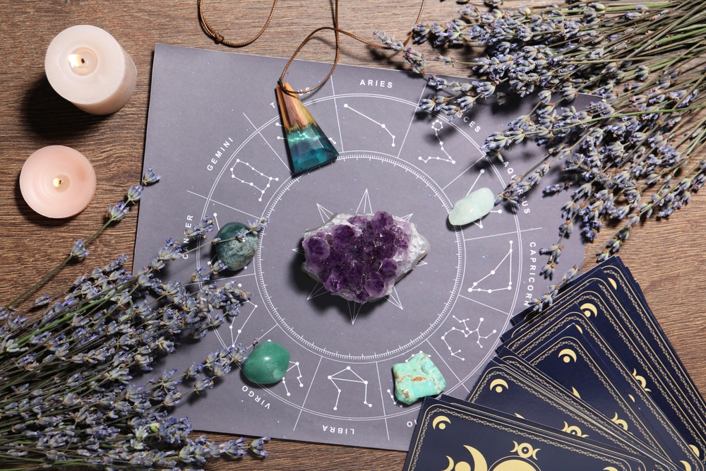 A collection of crystals arranged on an astrology mat displaying zodiac signs, surrounded by dried lavender and candles, with tarot cards placed nearby.